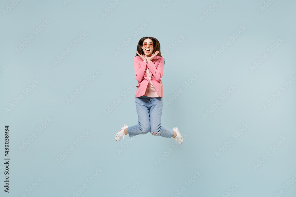 Full length young fun overjoyed excited happy caucasian trendy stylish woman 20s wearing pastel pink clothes glasses jump high hold face isolated on blue background studio. People lifestyle concept.