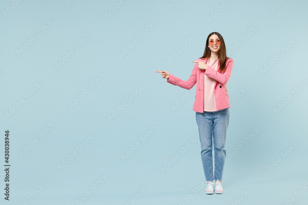 Full length young fun smiling trendy fashionable woman in pastel pink clothes glasses point index finger aside on workspace area isolated on blue background studio portrait. People lifestyle concept. Full length young fun smiling trendy fashionable woman in pastel pink clothes glasses point index finger aside on workspace area isolated on blue background studio portrait. People lifestyle concept.