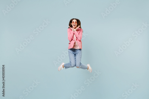 Full length young fun overjoyed excited happy caucasian trendy stylish woman 20s wearing pastel pink clothes glasses jump high hold face isolated on blue background studio. People lifestyle concept.
