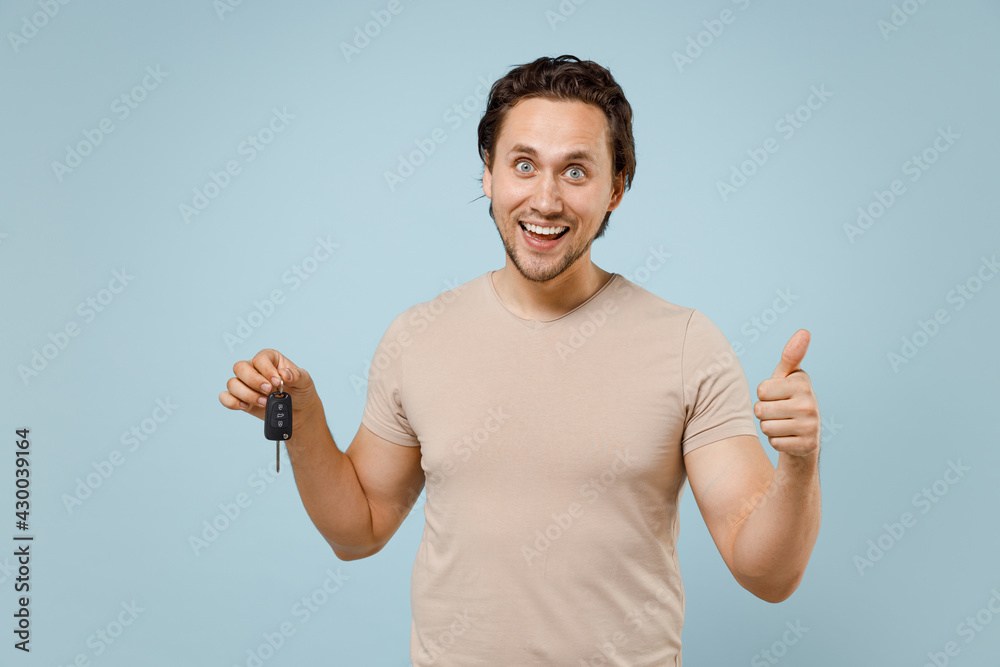 Young smiling happy satisfied caucasian man 20s wearing casual basic beige t-shirt holding car keys fob keyless system show thumb up like gesture isolated on pastel blue background studio portrait Young smiling happy satisfied caucasian man 20s wearing casual basic beige t-shirt holding car keys fob keyless system show thumb up like gesture isolated on pastel blue background studio portrait