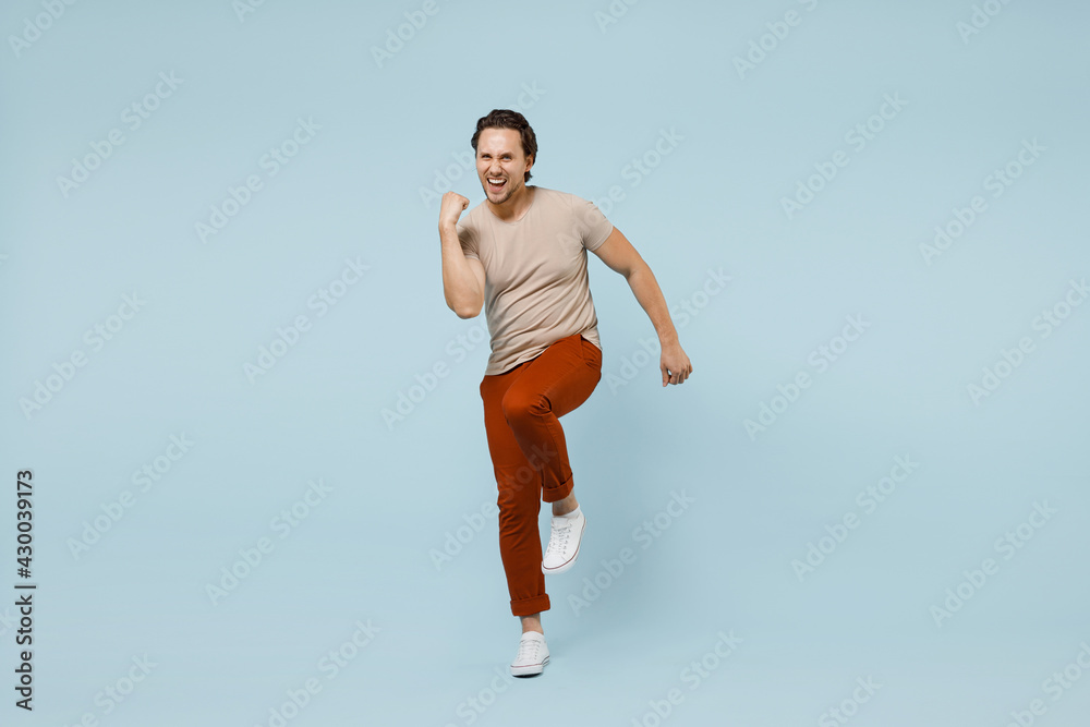 Full length young happy happy overjoyed excited man 20s wearing casual basic beige t-shirt do winner gesture clench fist with raised up leg isolated on pastel blue color background studio portrait