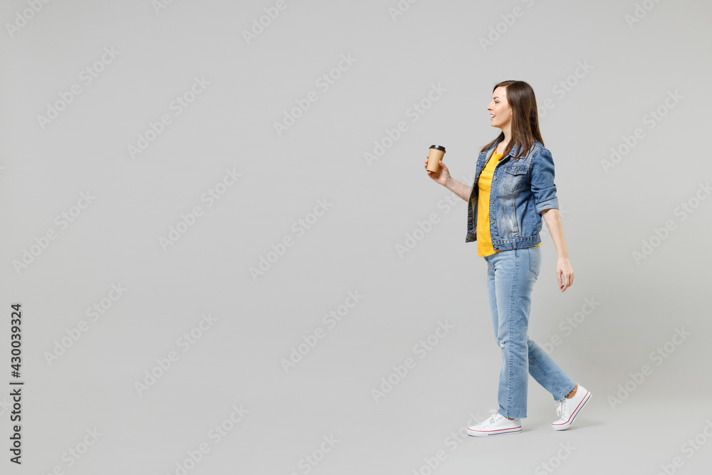Full length side view walk young happy caucasian woman in casual denim jacket yellow t-shirt holding in hand paper cup of coffee drink tea isolated on grey background studio. People lifestyle concept. Full length side view walk young happy caucasian woman in casual denim jacket yellow t-shirt holding in hand paper cup of coffee drink tea isolated on grey background studio. People lifestyle concept.