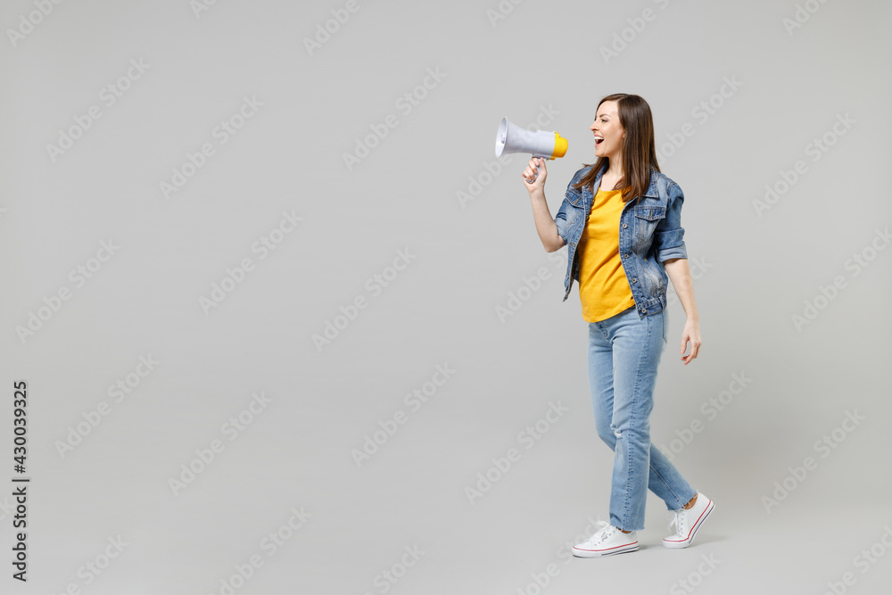 Side view full length young happy caucasian woman wearing casual trendy denim jacket yellow t-shirt screaming aside shouting in megaphone isolated on grey background studio. People lifestyle concept.