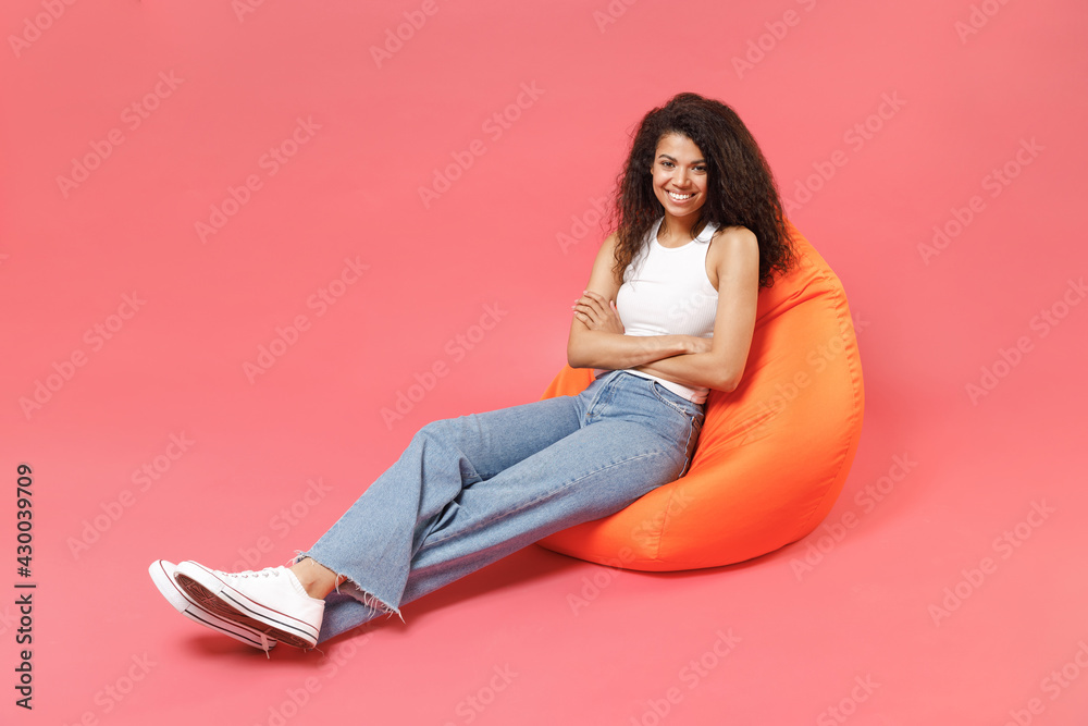Young fun smiling happy friendly positive cute african american woman 20s wearing casual white tank shirt sitting in orange bean bag chair hold hands crossed folded isolated on pink color background Young fun smiling happy friendly positive cute african american woman 20s wearing casual white tank shirt sitting in orange bean bag chair hold hands crossed folded isolated on pink color background