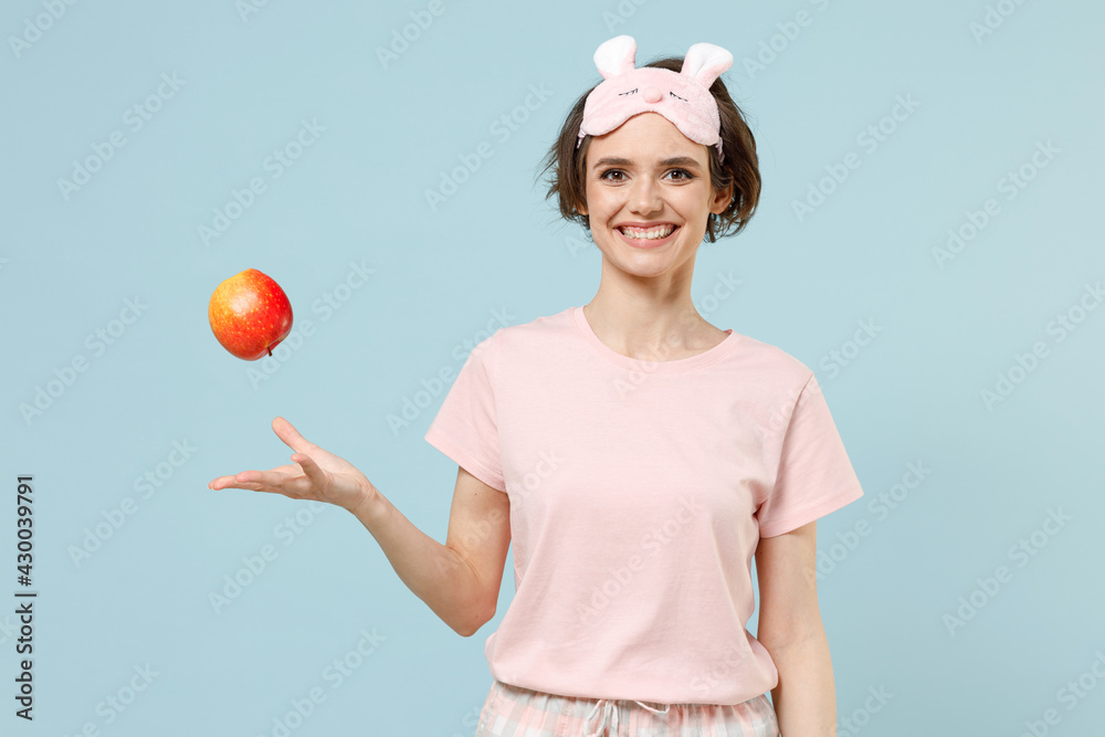 Young smiling happy fun woman wear pajamas jam sleep eye mask rest relaxing at home throws up red apple fruit isolated on pastel blue background studio. Healthy vegeterian food night bedtime concept. Young smiling happy fun woman wear pajamas jam sleep eye mask rest relaxing at home throws up red apple fruit isolated on pastel blue background studio. Healthy vegeterian food night bedtime concept.