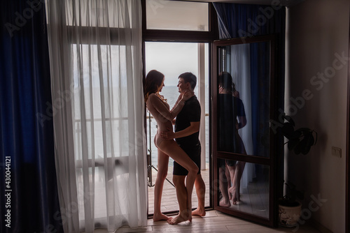 Silhouettes of young loving couple in front of panoramic window overlooking the sea. Lovers travel on their honeymoon, spend a date in hotel room. Beautiful woman hugs a man, kiss, look at each other