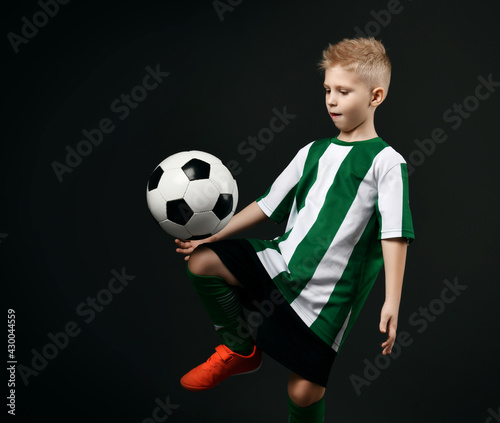 Blond boy child in sports green and white clothes playing soccer ball game over dark background. Trendy sports active children fashion concept © Dmitry Lobanov