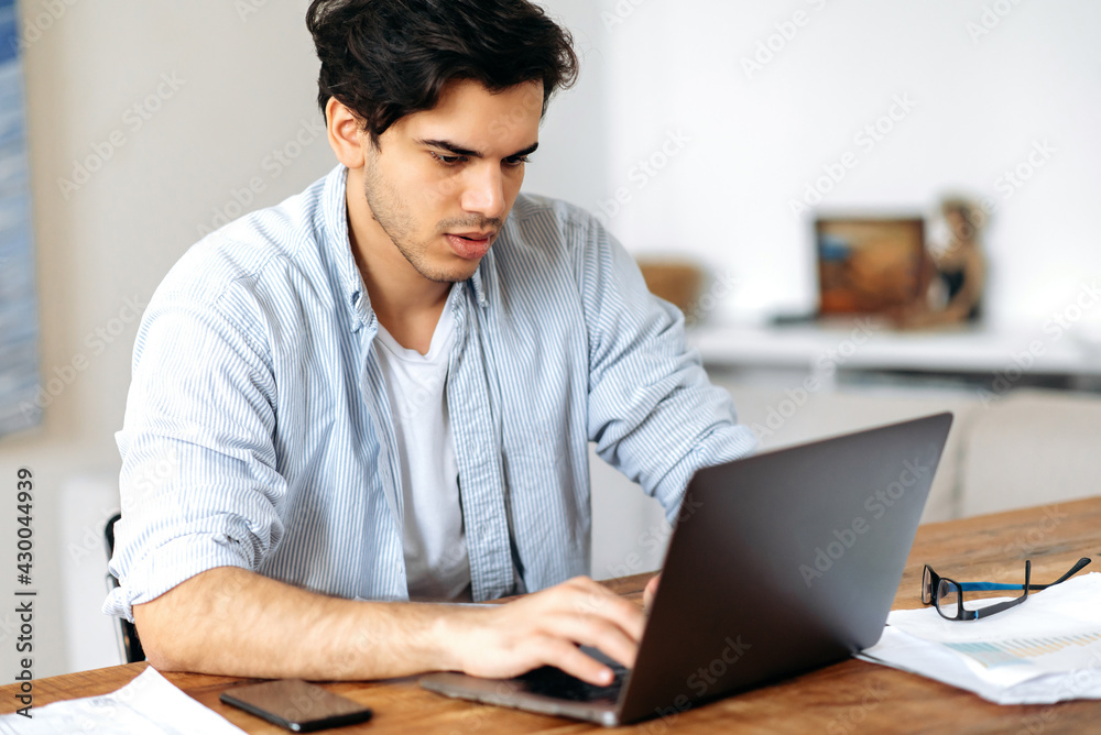 Young serious successful mixed race man, freelancer, manager or student, working or studying remotely, in casual wear, uses laptop, texting with colleague or client, browsing internet, searching ideas Young serious successful mixed race man, freelancer, manager or student, working or studying remotely, in casual wear, uses laptop, texting with colleague or client, browsing internet, searching ideas