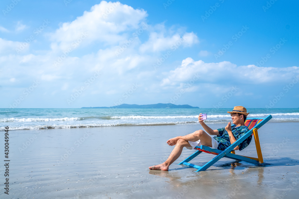 Young Asian man resting on sunbed on tropical beach. Happy guy sitting on beach  chair by