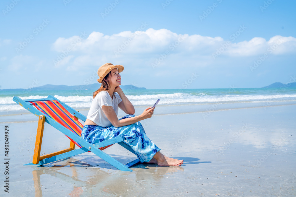 Asian woman resting on sunbed on the beach. Happy girl sitting on beach chair by the sea using smartphone for online shopping or text message. Female enjoy beach outdoor lifestyle on summer vacation