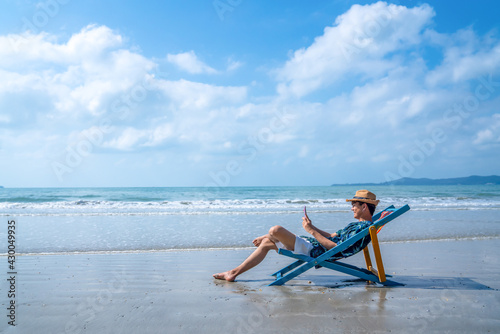 Asian man resting on sunbed on tropical beach. Happy guy sitting on beach chair by the sea using smartphone for selfie or video call. Handsome male enjoy beach outdoor lifestyle on summer vacation