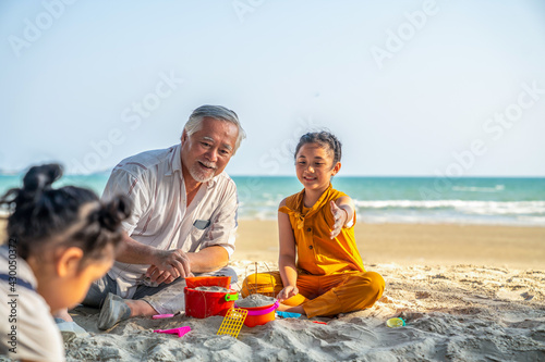 Happy Asian family on summer travel vacation. Grandfather enjoy play beach toys with two grandchild girl on sand beach in sunny day. Senior man with granddaughter having fun outdoor lifestyle activity