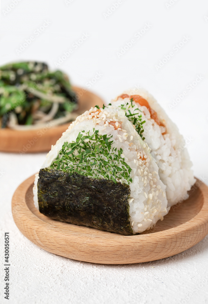 Two japanese onigiri triangled rice balls with salmon, seasoning, roasted nori and sesame seeds on a wooden plate and on white background Two japanese onigiri triangled rice balls with salmon, seasoning, roasted nori and sesame seeds on a wooden plate and on white background