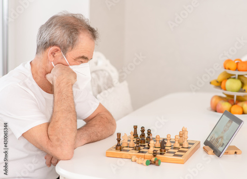 Pensive senior man wearing protective face mask uses tablet computer to play chess with his friends during quarantine Coronavirus (Covid-19) epidemic photo