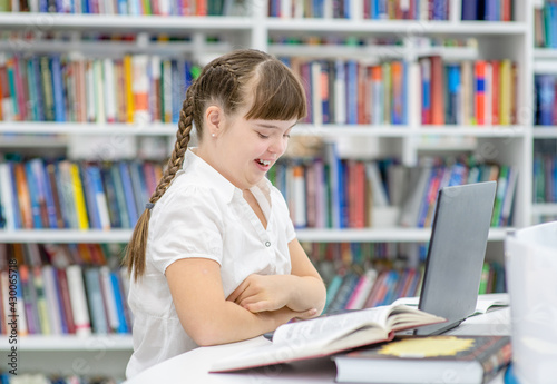 Smiling young girl with syndrome down uses a laptop at library. Education for disabled children concept