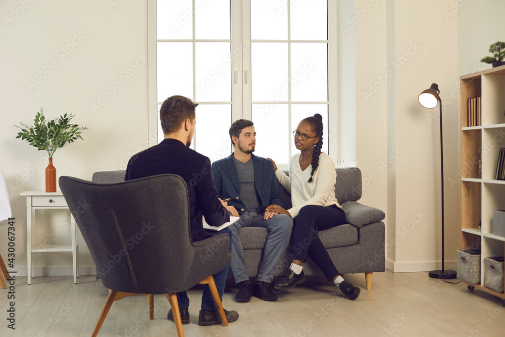Therapy session. Young couple asking family psychologist to help save their marriage. Multicultural boyfriend and girlfriend sitting on couch discussing relationship issues and problems with therapist