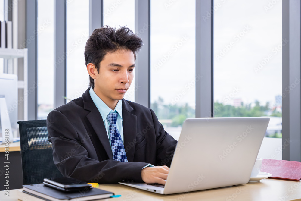 Young Asian man sitting at office desk and working with laptop computer. Businessman or salary man life.