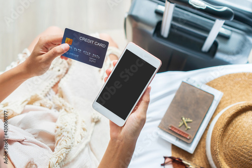 Happy traveler Asian woman holding credit card while using mobile phone with travel accessories on bed. Online shopping and travel, cashless society concept