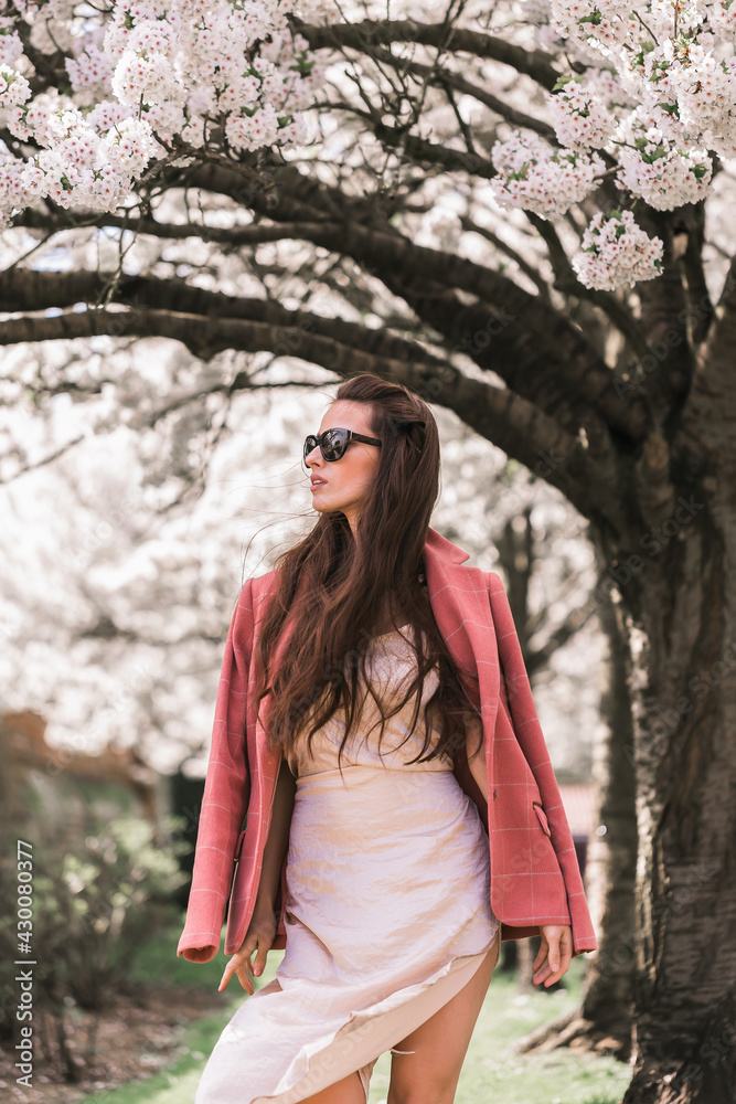 Beautiful young brunette girl, model, in a fashionable dress and a classic checkered pink jacket. Sunglasses. Looks away. Background of flowering trees.