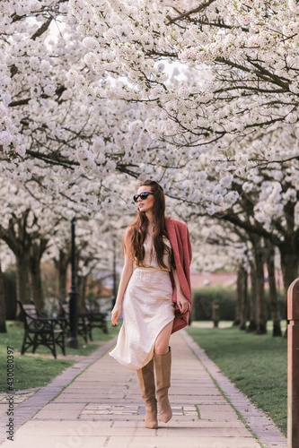 Beautiful young brunette girl, model, in a fashionable dress and a classic checkered pink jacket. Sunglasses. In motion. Looking away. Background of the alley of flowering trees.