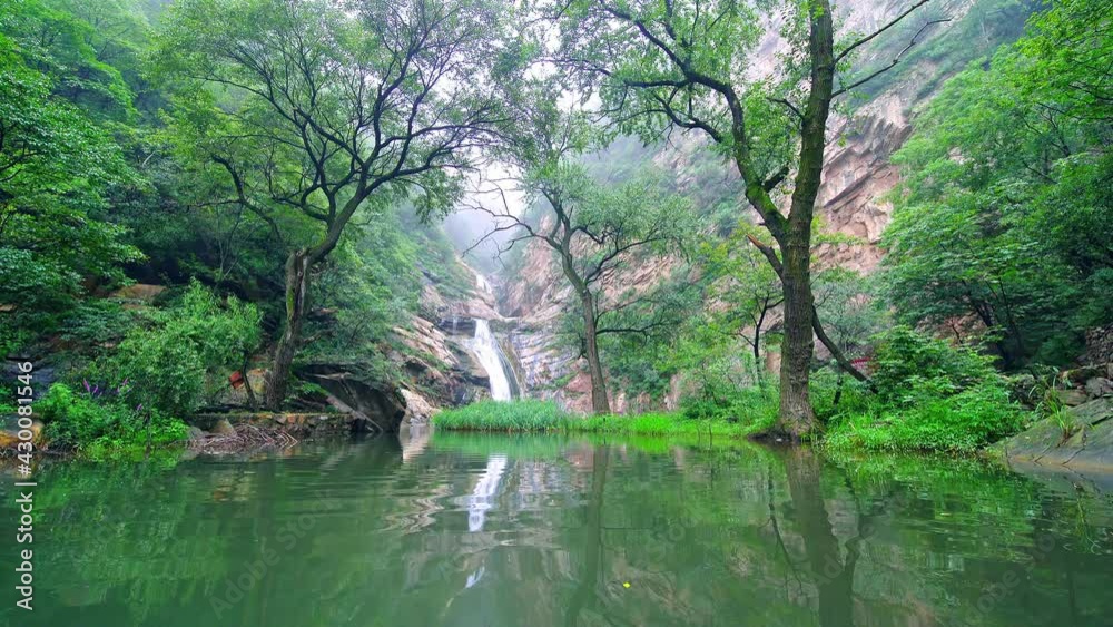 In gorge, a distant stream flowing from mountains flows into a creek. The reflection of surrounding green trees in clear river water In gorge, a distant stream flowing from mountains flows into a creek. The reflection of surrounding green trees in clear river water