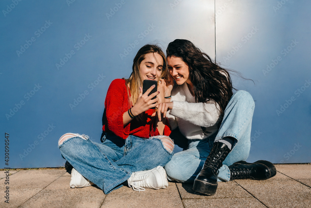 Couple of friends with long hair, one blonde and one brunette, looking at their cell phones sitting on the floor in front of a blue wall. Couple of friends with long hair, one blonde and one brunette, looking at their cell phones sitting on the floor in front of a blue wall.