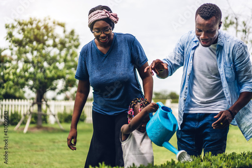 Portrait of enjoy happy love black family african american father and mother with little african girl child smiling and watering garden grass with rubber strap in summer park at home