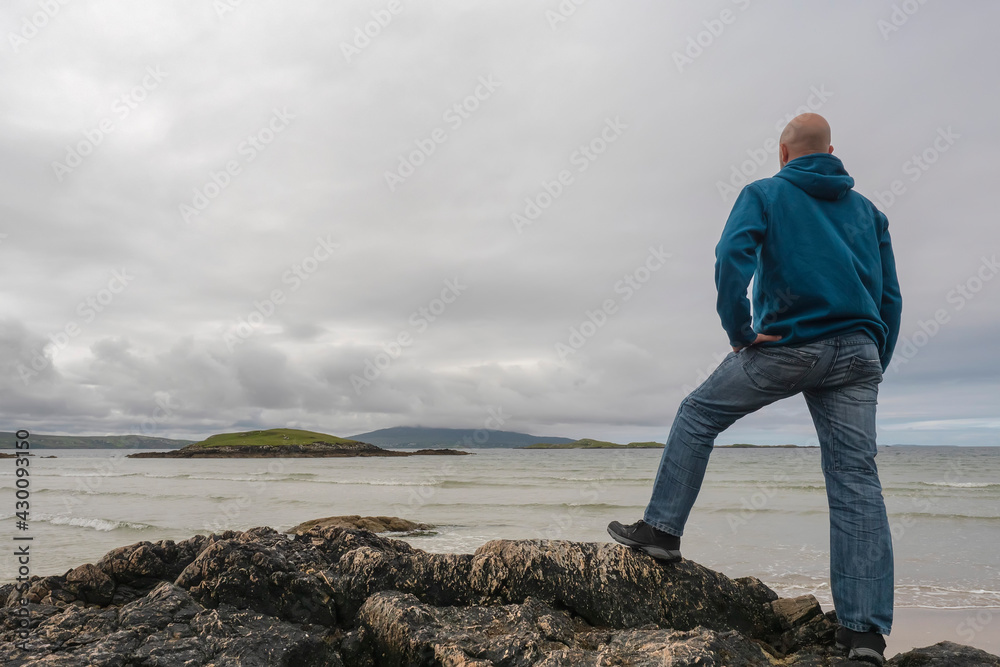 Portrait of bald man standing on a rock by the ocean, hands on his hips, back to camera, beautiful and calm scenery in the background. West coast of Ireland. Travel and tourism concept