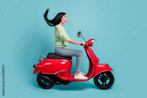 Profile side view of attractive cheerful amazed girl riding moped fast speed air blowing hair isolated over bright blue color background © deagreez