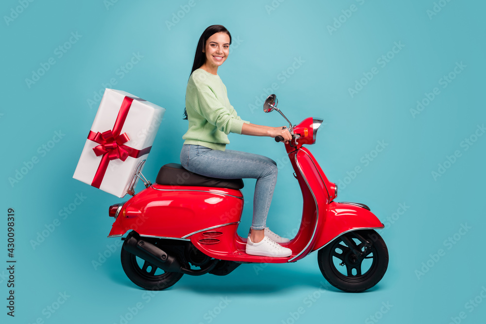 Profile side view of attractive cheerful girl riding moped delivering giftbox holiday isolated over bright blue color background Profile side view of attractive cheerful girl riding moped delivering giftbox holiday isolated over bright blue color background