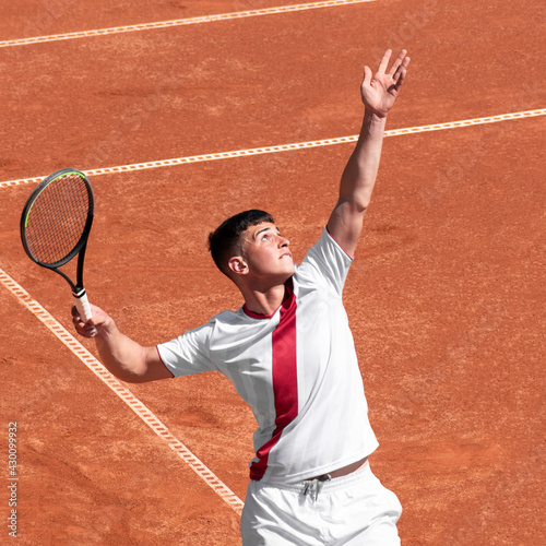 Young male tennis player performs serves ball on clay tennis court at start of game. Cute man athlete in action. Individual, competitive sport © Elena
