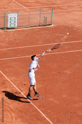 Young tennis player jump and hit ball while serving on orange clay court at start of match. Athlete in motion during game. Vertical sports background, banner, copy space © Elena