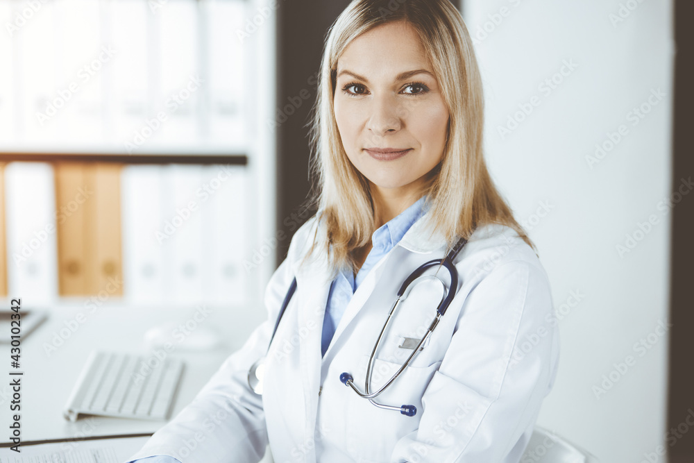 Friendly female doctor sitting in clinic at her working place. Portrait of cheerful smiling physician. Perfect medical service in hospital. Medicine and healthcare concept Friendly female doctor sitting in clinic at her working place. Portrait of cheerful smiling physician. Perfect medical service in hospital. Medicine and healthcare concept