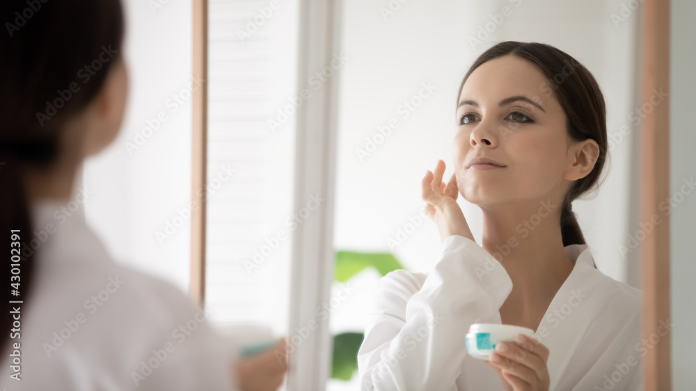 Satisfied cosmetic product consumer applying cream, looking in mirror, touching perfect facial skin. Young woman doing skincare moisturizing procedure after taking bath. Mirror reflection head shot