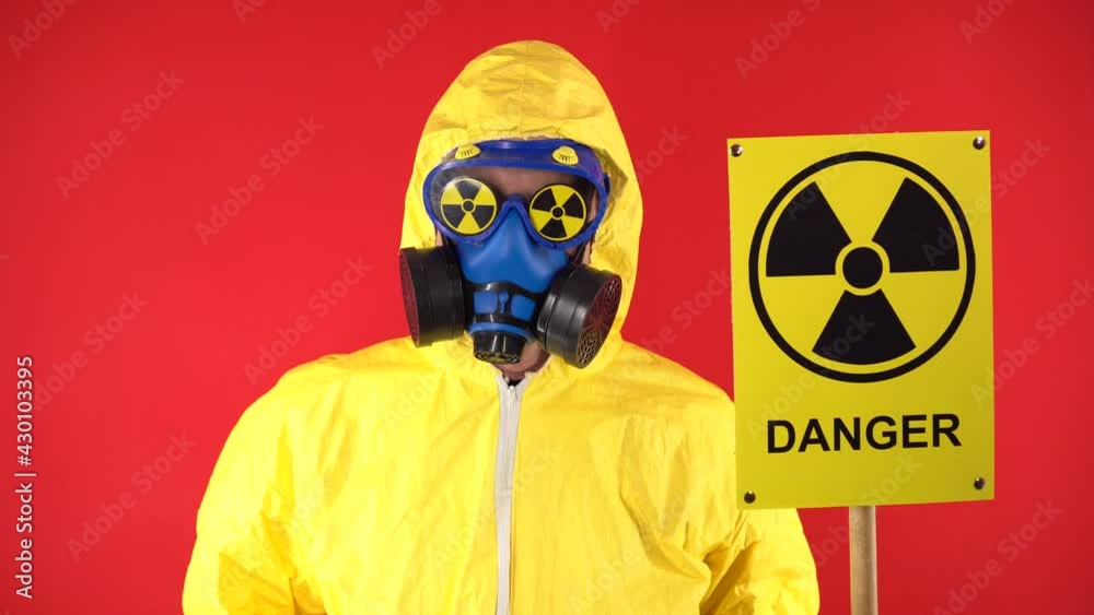 A man in a protective chemical suit shakes his head negatively with a poster of Radiation. Isolate, studio shooting. Radiation hazard concept on red background A man in a protective chemical suit shakes his head negatively with a poster of Radiation. Isolate, studio shooting. Radiation hazard concept on red background