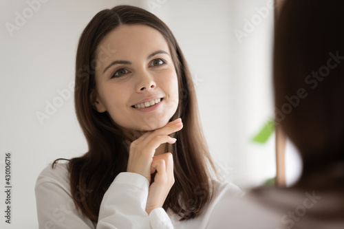 Happy beautiful teen girl looking in mirror, admiring reflection , skincare moisturizing and cleansing cream effect, touching perfect smooth facial skin, looking at herself, smiling. Head shot