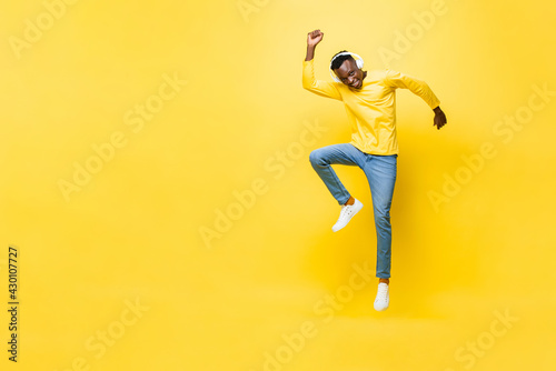 Happy energetic young African man wearing headphones listening to music and jumping with hand up next to empty space in yellow isolated studio background