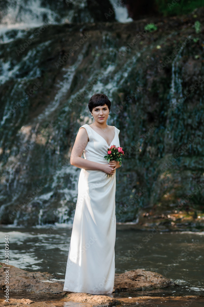 A girl in a white dress stands against the background of a waterfall. The bride stands against the background of a waterfall