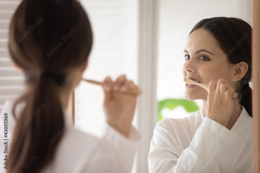 Young woman in bathrobe brushing teeth with wooden bamboo toothbrush. Teen girl choosing eco friendly tools for morning bath routine, oral mouth hygiene, protection from caries. Mirror reflection Young woman in bathrobe brushing teeth with wooden bamboo toothbrush. Teen girl choosing eco friendly tools for morning bath routine, oral mouth hygiene, protection from caries. Mirror reflection