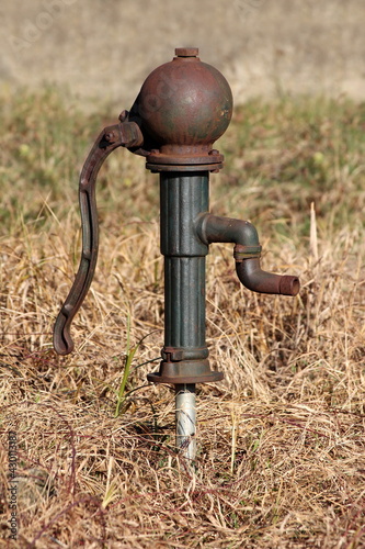 Strange and unusually shaped vintage retro partially rusted wrought iron dark green hand water pump mounted on grey water pipe surrounded with dry grass in suburban family house backyard on warm sunny