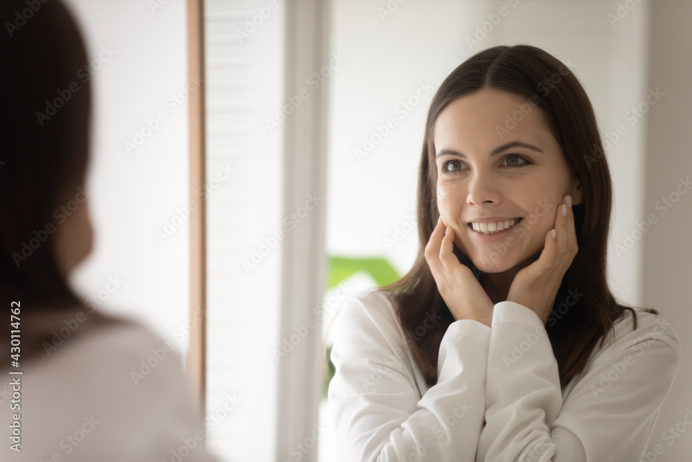 Happy pretty young woman wearing bathrobe, touching perfect smooth pure facial skin on cheeks, looking in mirror, smiling at reflection. Teen girl excited about home spa cosmetic effect. Head shot