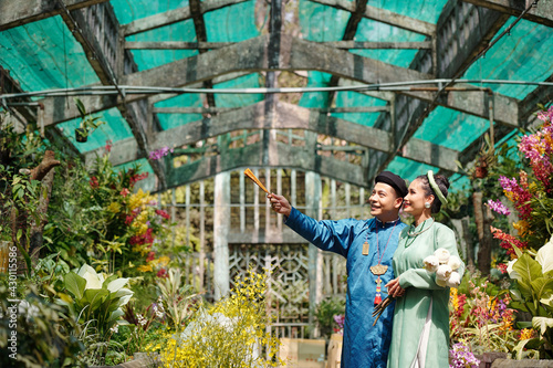 Happy young Vietnamese man showing beautiful flowers in greenhouse to bride