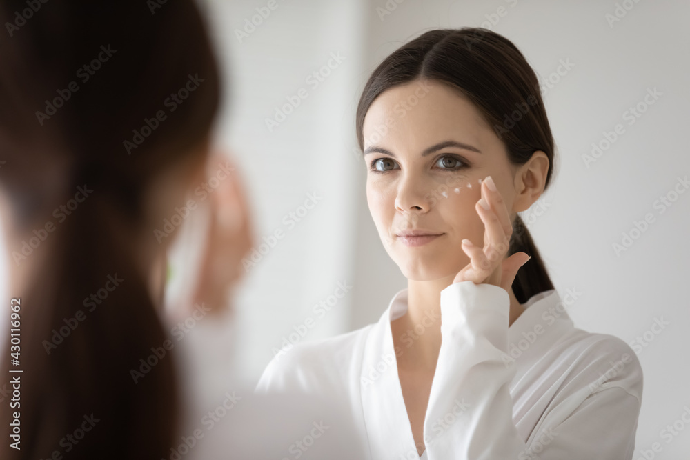 Mirror reflection of girl cleaning and moisturizing facial skin with lotion. Young woman in bathrobe taking spa skincare procedures at home, applying cream, touching face with finger. Head shot Mirror reflection of girl cleaning and moisturizing facial skin with lotion. Young woman in bathrobe taking spa skincare procedures at home, applying cream, touching face with finger. Head shot