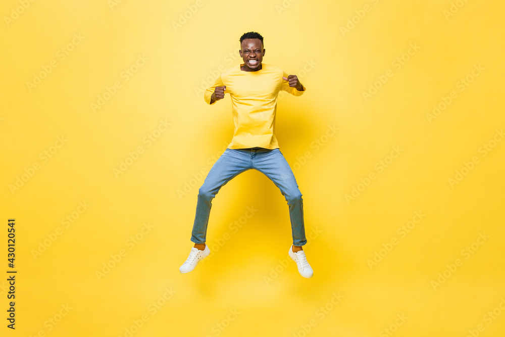 Young energetic excited African man jumping and clenching hands on isolated yellow studio background