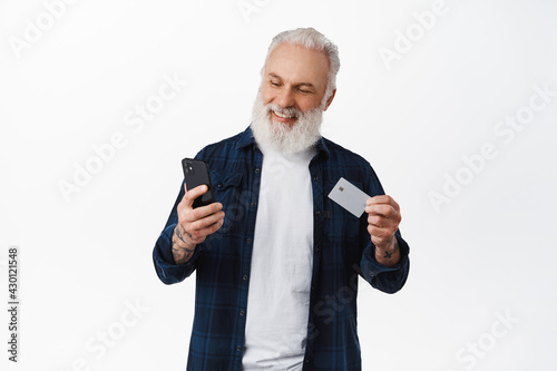Handsome mature man paying with credit card on smartphone, showing card and looking at phone with smiling face, shopping online, standing against white background