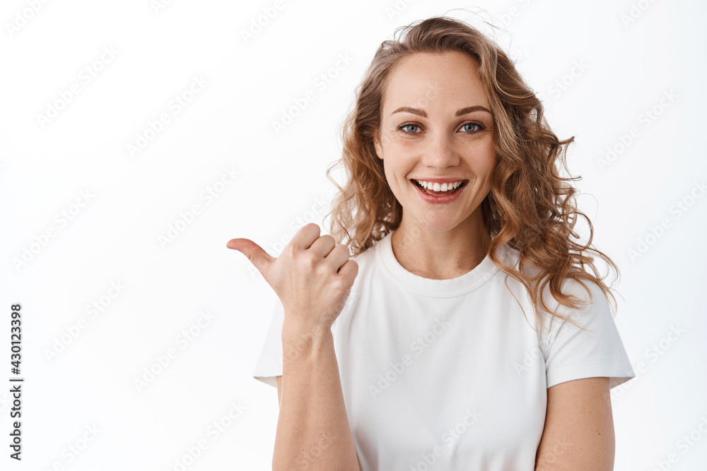 Image of stylish blond woman points left, shows promotional text aside, smiles and looks satisfied at camera, white background Image of stylish blond woman points left, shows promotional text aside, smiles and looks satisfied at camera, white background