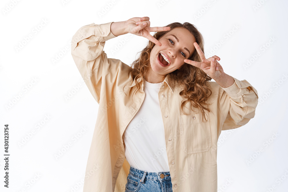 Fototapeta premium Positive blond woman showing peace v-signs on face, smiling and looking happy, having fun, standing over white background