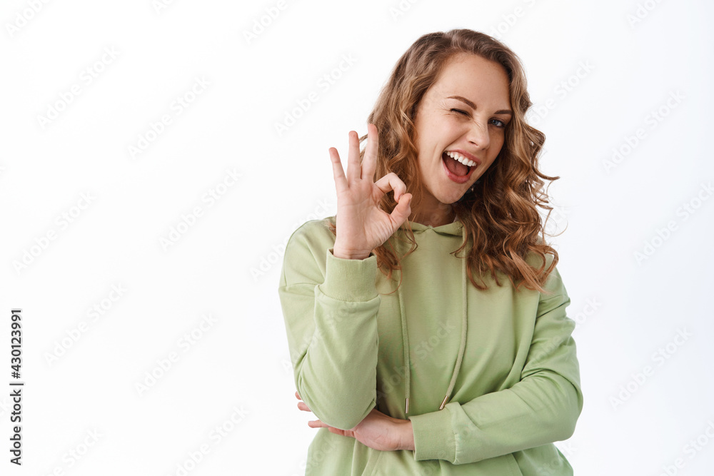Positive blond girl winking, showing okay sign in approval, say yes, agree and like, praise nice work, good choice, standing over white background Positive blond girl winking, showing okay sign in approval, say yes, agree and like, praise nice work, good choice, standing over white background