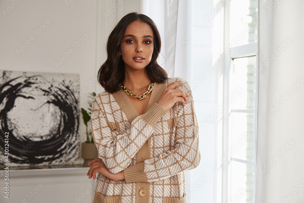 Pretty fashion beautiful woman sexy lady brunette curly hair dark tanned skin wear trend clothes knitted beige suit jacket top pants shoes interior room sofa plants spring collection luxury.