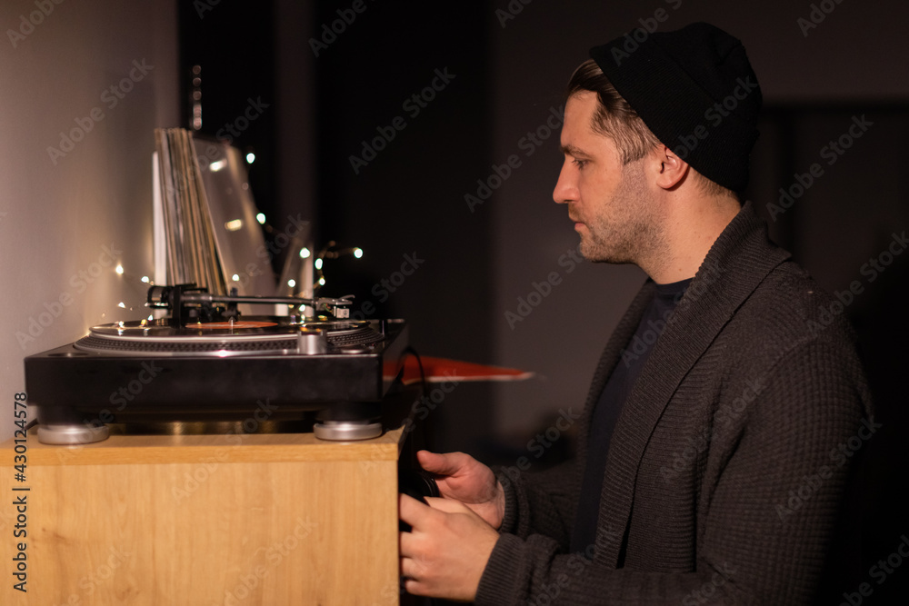 Young unshaven handsome millennial guy puts record on turntable and taking headphones. Musical Hobby Concept. anti-digital hobby In stylish home environment